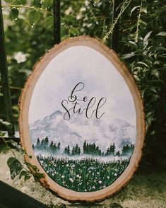 a wood slice with the words be still painted on it and mountains in the background