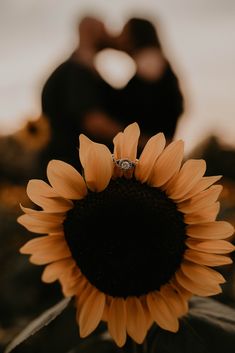 an engaged couple kissing in front of a sunflower with their engagement ring on it