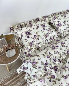 a bed with purple flowers on it next to a night stand and side table in the corner