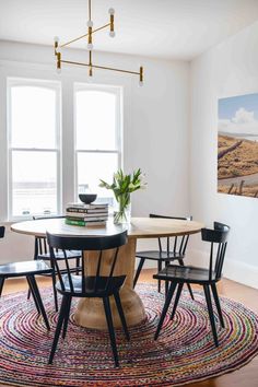 a dining room table with four chairs and a rug on the floor in front of two windows