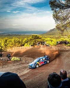 a rally car driving down a dirt road in front of a group of people watching