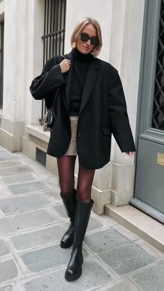 a woman walking down the street wearing black boots and a blazer over her shoulders