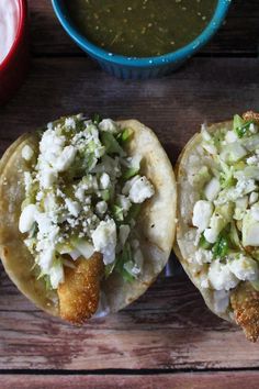 two tacos with meat, cheese and sauces on the side sitting on a wooden table