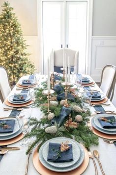 a dining room table set for christmas with blue and gold place settings, silverware, candlesticks and greenery