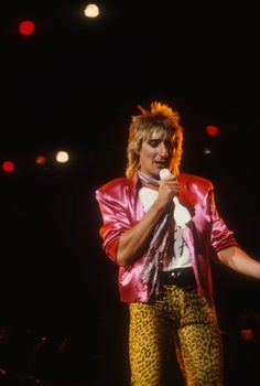 a man in pink jacket and leopard print pants on stage holding a microphone with his right hand