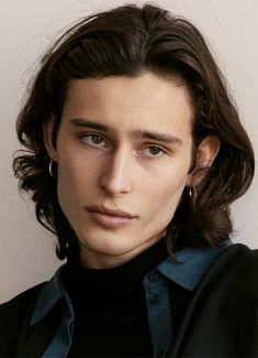 a young man with long hair wearing a black suit and blue turtle neck shirt looking at the camera