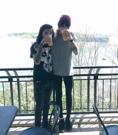 two people standing next to each other on a balcony with water and trees in the background