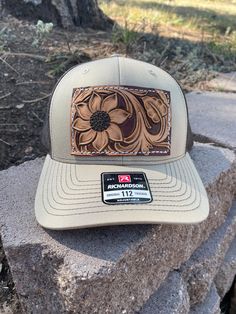 A tan and brown Richardson Trucker hat decked out with a sunflower and some vines on a leather patch! These Richardson  hats are super comfortable and have an adjustable snap back.  PLEASE READ: This item is for pre-ordering, and is made to order! Once purchased, this item will take approximately two weeks to re-create. Item will be a close replica, but each one will have slight variances in color, carving, etc. as I am not perfect, and leather is not perfect either! *All of HCL's leather products are cut, designed, dyed, painted, sewn, and assembled by hand. Items will not be machine perfect, but I try my hardest to get them there! Please keep in mind that leather may have natural scuffs, brand marks, discoloration, etc. It's all part of what makes leather so beautiful and unique! Brown Rodeo Hat With Leather Patch, Western Brown Trucker Hat With Flat Bill, Western Style Brown Trucker Hat With Flat Bill, Brown Trucker Hat For Western-themed Events, Brown Flat Bill Hat For Western-themed Events, Adjustable Brown Brimmed Trucker Hat, Brown Western Style Baseball Cap, Adjustable Brown Trucker Hat For Country Events, Brown Adjustable Trucker Hat For Country Events