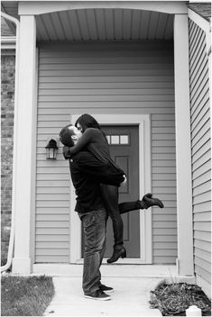 a man and woman standing in front of a house hugging each other with their arms around one another