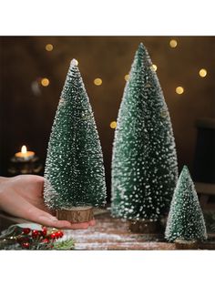 three small christmas trees sitting on top of a wooden table next to a person's hand