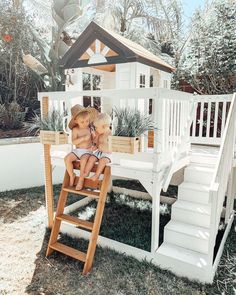 two children are sitting on a wooden chair in front of a tree house and stairs