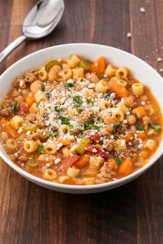 a white bowl filled with pasta and meat soup on top of a wooden table next to a spoon