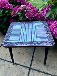 a mosaic table sitting on top of a sidewalk next to some purple hydrangeas