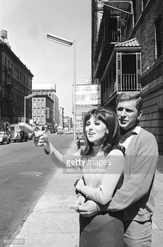 a man and woman standing next to each other on the sidewalk in front of tall buildings