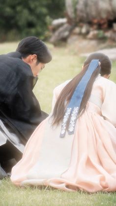 two people sitting in the grass with one woman wearing a long dress and another man standing behind her