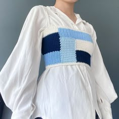 a woman wearing a white shirt with blue squares on the chest and sleeves, standing in front of a gray wall