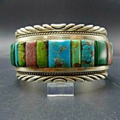 "PHILLIP MORSE TURQUOISE BRACELET DESCRIPTION: This remarkable bracelet is from trending Dine'/Chippewa silversmith Phillip Morse. Taught by his legendary Aunt, Verna Blackgoat, Phillip puts in long hours to ensure perfection in his silverwork, and uses only the finest natural gemstones. The perfectly arranged cobblestone inlay boasts an assortment of gemstones, including Broken Arrow, Fox, Royston, and Kingman turquoise, green malachite, blue lapis, and purple lepidolite. The gemstones are secu Southwestern Bangle Cuff Bracelet With Natural Stones, Southwestern Style Natural Stones Cuff Bangle, Multicolor Handmade Cuff Bracelet Collectible, Traditional Turquoise Bracelet With Inlay, Handmade Multicolor Cuff Bracelet Collectible, Artisan Multi-stone Bangle Bracelet, Multicolor Natural Stones Cuff Bangle, Turquoise Inlay Bracelet For Collectors, Southwestern Turquoise Inlay Bracelet