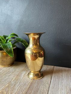 two gold vases sitting on top of a wooden table next to a green plant