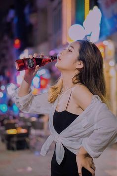 a woman standing on the street drinking from a coca - cola bottle in front of neon lights