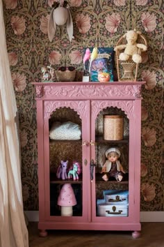 a pink cabinet with stuffed animals and toys on it's shelves in front of a floral wallpapered wall