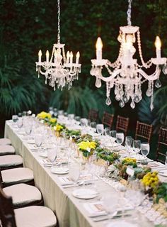 a long table is set with white plates and yellow flowers, chandeliers are hanging from the ceiling