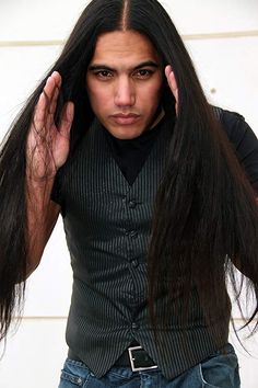 a man with long black hair holding his hands up to his ears and looking at the camera