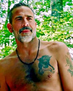 a man with tattoos on his chest standing in front of some trees and bushes, looking off to the side