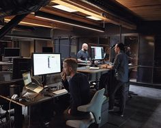 three men are working on computers in an office setting with wood paneling and exposed ceilings