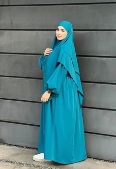 a woman wearing a blue hijab stands against a wall with her hands on her hips