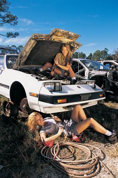 two women sitting on the ground next to a car with its hood open and hoses in front of it