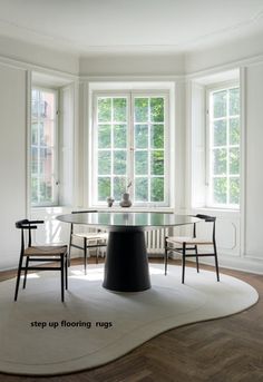 a round table with chairs around it in front of two windows and a rug on the floor