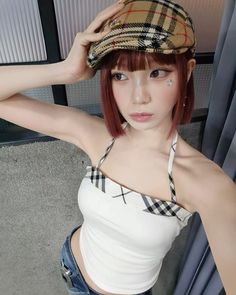 a woman with red hair wearing a white tank top and plaid cap, standing in front of a window