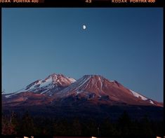 the moon is setting behind a mountain with snow on it's top and trees below