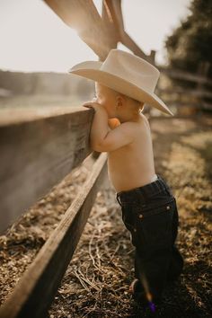 a small child wearing a cowboy hat leaning against a fence with his hands on the side