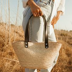 Easily pack up the Jude tote in a suitcase so you can enjoy its versatility with the outfits you've packed for your travels. Its unique shape, with neutral design elements make it the bag that goes with everything. Each handmade basket is unique and may have small imperfections or vary in size and color. All Leah products are responsibly sourced and ethically crafted. Handles: Double layer black leather handles No interior lining Length: 9" Width: 16" Height: 8" Handle Drop: 10" Imported Chic Natural Beach Bag With Handles, Chic Natural Color Bucket Bags, Modern Bucket Bag For Vacation, Chic Beige Basket Shoulder Bag, Casual Basket Bag With Braided Handles, Casual Beige Basket Bag, Chic Natural Color Bucket Shoulder Bag, Chic Canvas Beach Bag For Travel, Casual Natural Shoulder Bag With Leather Handles