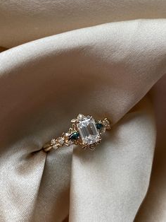 an engagement ring is sitting on top of a white satin material with gold accents and green stones