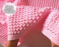 a pink knitted blanket laying on top of a white tablecloth next to a person's hand