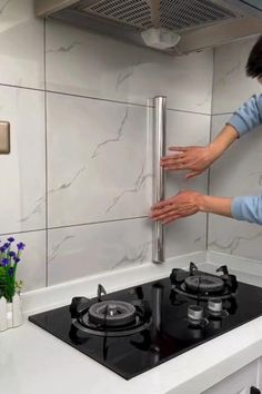 a woman reaching for something on top of a gas stove in a white tiled kitchen