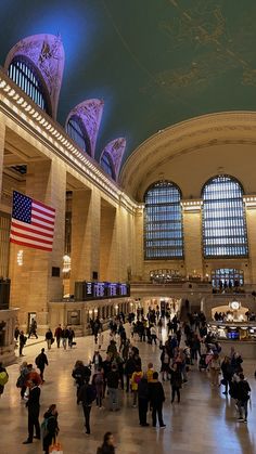 many people are walking around in the lobby of a building with large windows and an american flag hanging from the ceiling