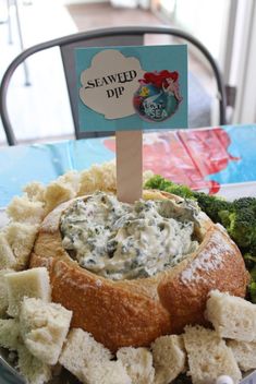 a bowl filled with dip surrounded by crackers and broccoli on a table