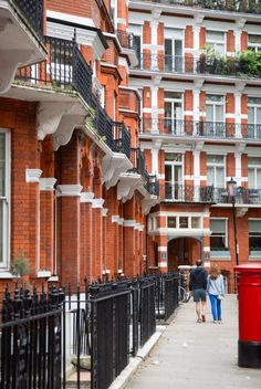 two people walking down the sidewalk in front of some brick buildings with balconies