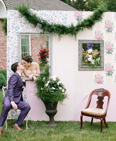 a man and woman kissing in front of a house with flowers on the wall behind them