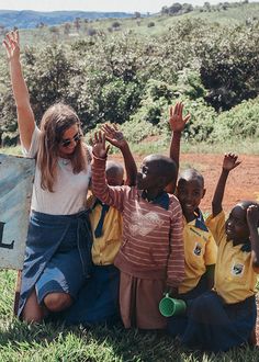 a group of children sitting on the ground with their hands in the air and one woman standing behind them