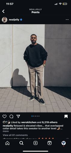 a man standing in front of a white wall wearing a black sweater and khaki pants