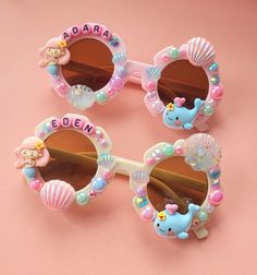 three children's sunglasses with hello kitty and mermaid decorations on them, sitting on a pink surface