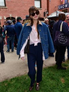 a woman wearing sunglasses and a denim jacket is standing on the grass in front of a crowd