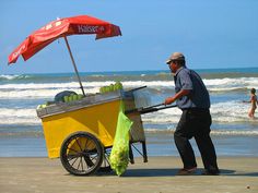 a man pushing a cart filled with fruit on top of a beach next to the ocean