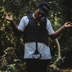 a man wearing a black vest and hat standing in the woods with his hands out