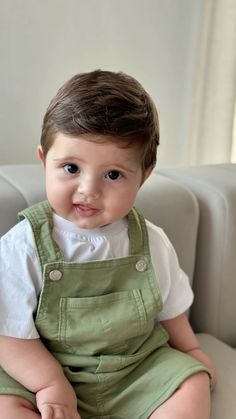 a little boy sitting on top of a couch wearing green overalls and smiling at the camera