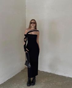 a woman standing in front of a white wall wearing a black dress with one shoulder cut out